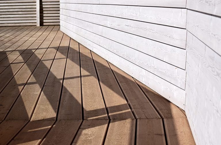 Composite decking longevity can be up to 30 years with today's performance-based materials and technology. A close-up view of composite decking against a house's white wood siding.